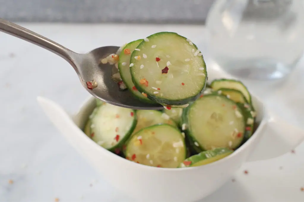 2 cucumber kimchi rounds being held up on a spoon over a white dish of cucumber kimchi on a grey and white marble surface