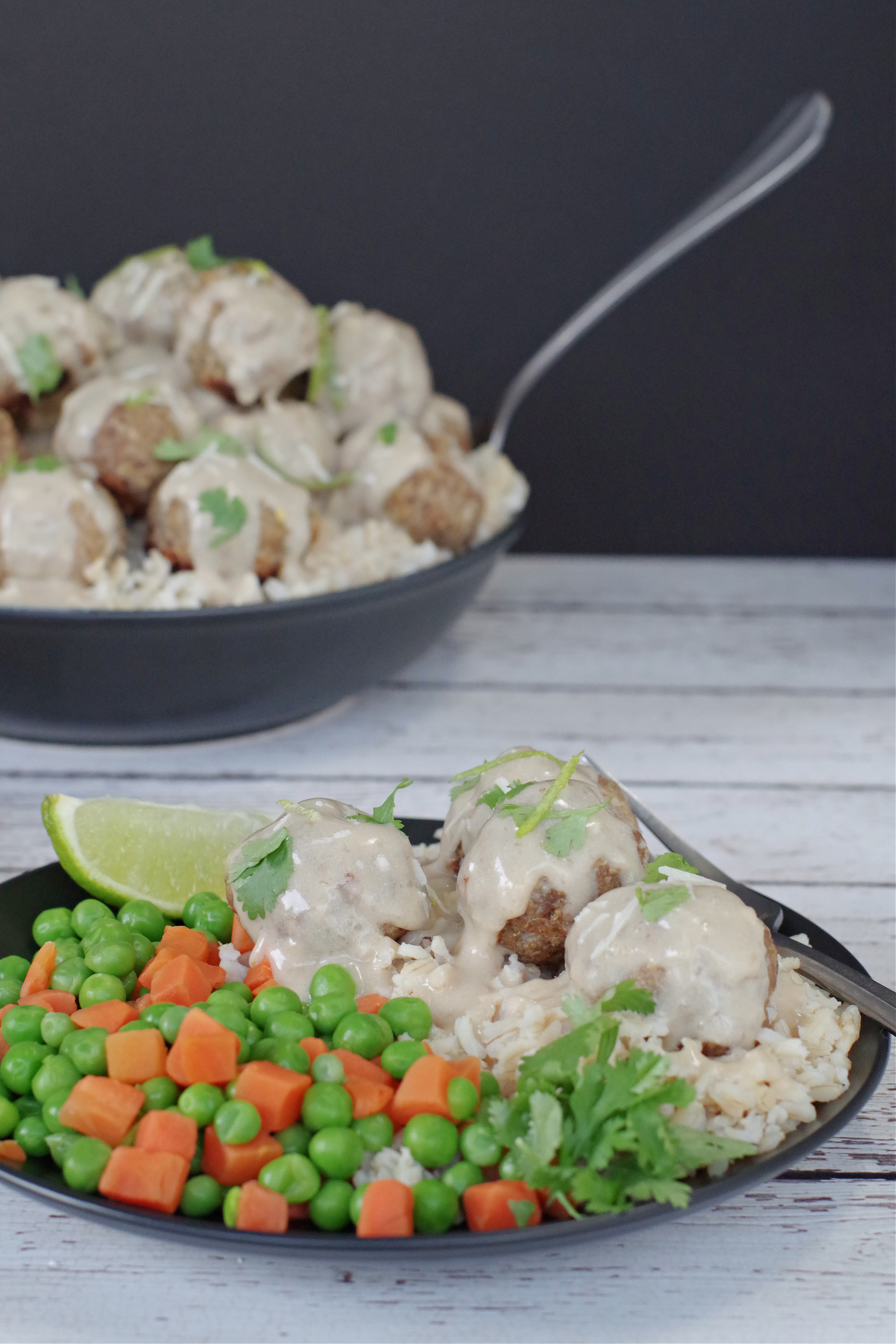 Healthy turkey meatballs with sauce on black plate, with veggies and rice and black bowl with turkey meatballs with white sauce in background