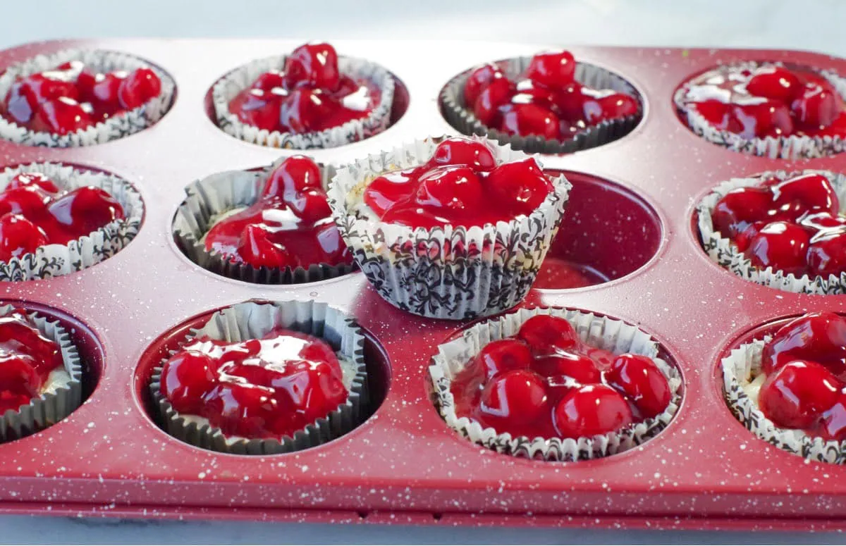 mini cherry cheesecakes in a red speckled muffin tin, with 1 cheesecake sitting on top