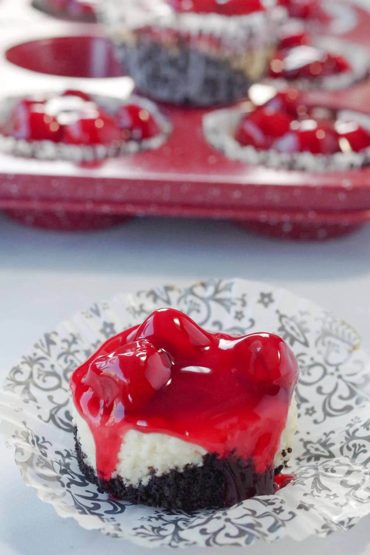 a mini cherry cheesecake on a damask print muffin wrapper, with a red speckled muffin tin ofmini cherry cheesecakes in the background
