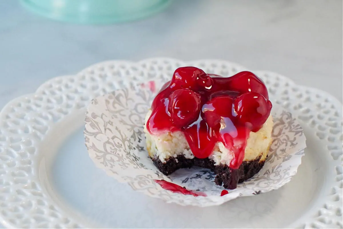 A mini cherry cheesecake with a bit taken out of it, on an open black and white damask muffin liner, sitting on a white (lattice edged) plate.