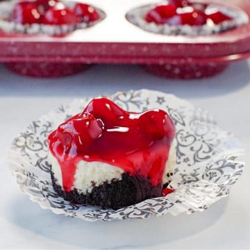 a mini cherry cheesecake on a damask print muffin wrapper, with a red speckled muffin tin ofmini cherry cheesecakes in the background