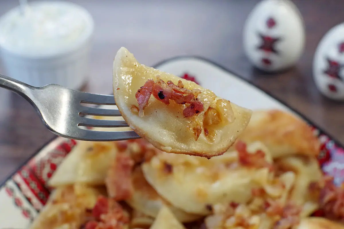 potato and cheese pierogi being held up on a fork over a dish of perogies with bacon and onion with sour cream and ukrainian print salt and pepper shakers in the background