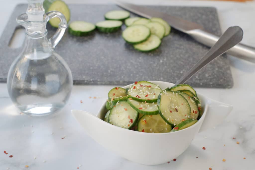 quick Kimchi on grey marble counter surface with glass vinegar container in background as well as a grey cutting board with sliced cucumbers and a knife