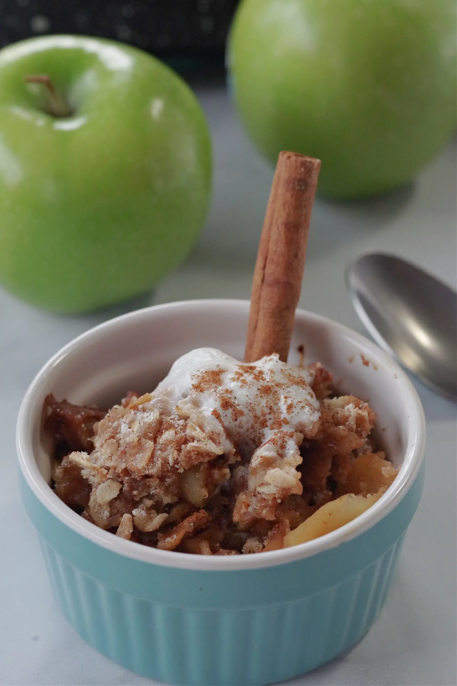 weight watchers apple crisp in a small blue bowl with a cinnamon stick in it and 2 green apples and a spoon in the background