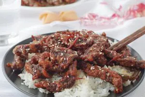 ginger beef over rice on a black plate on white table cloth, with fortune cookies