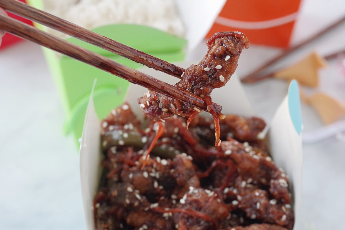 a piece of ginger beef being held up with chopsticks over a blue take-out container