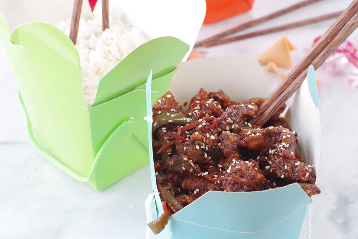 ginger beef in blue take-out container with chopsticks and green take-out container with rice in the background