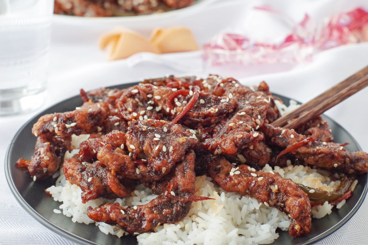 ginger beef over rice on a black plate on white table cloth, with fortune cookies