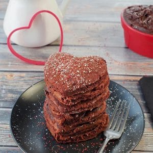stack of heart-shaped red velvet pancakes on a black plate with a heart cookie cutter in the background
