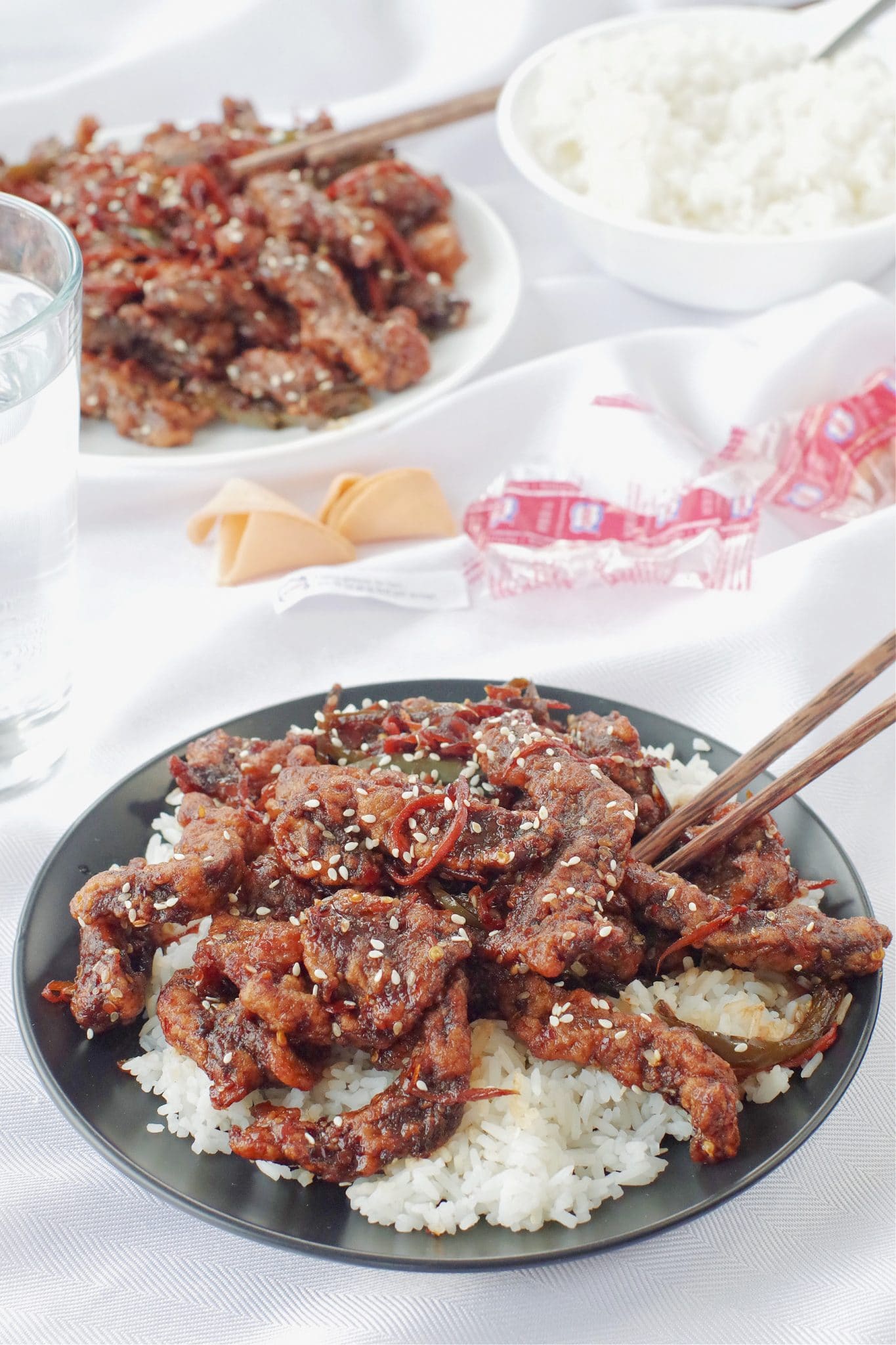 ginger beef over rice on black plate, with chopsticks on a white tablecloth, with a dish of ginger beef and rice in the background, as well as fortune cookies