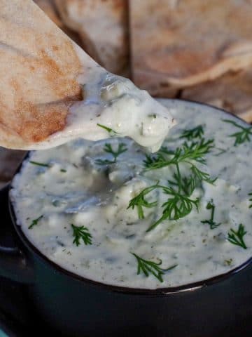 triangle of pita bread being dipped into tzatziki sauce in black bowl on black tray with pita bread in background and lemon wedges