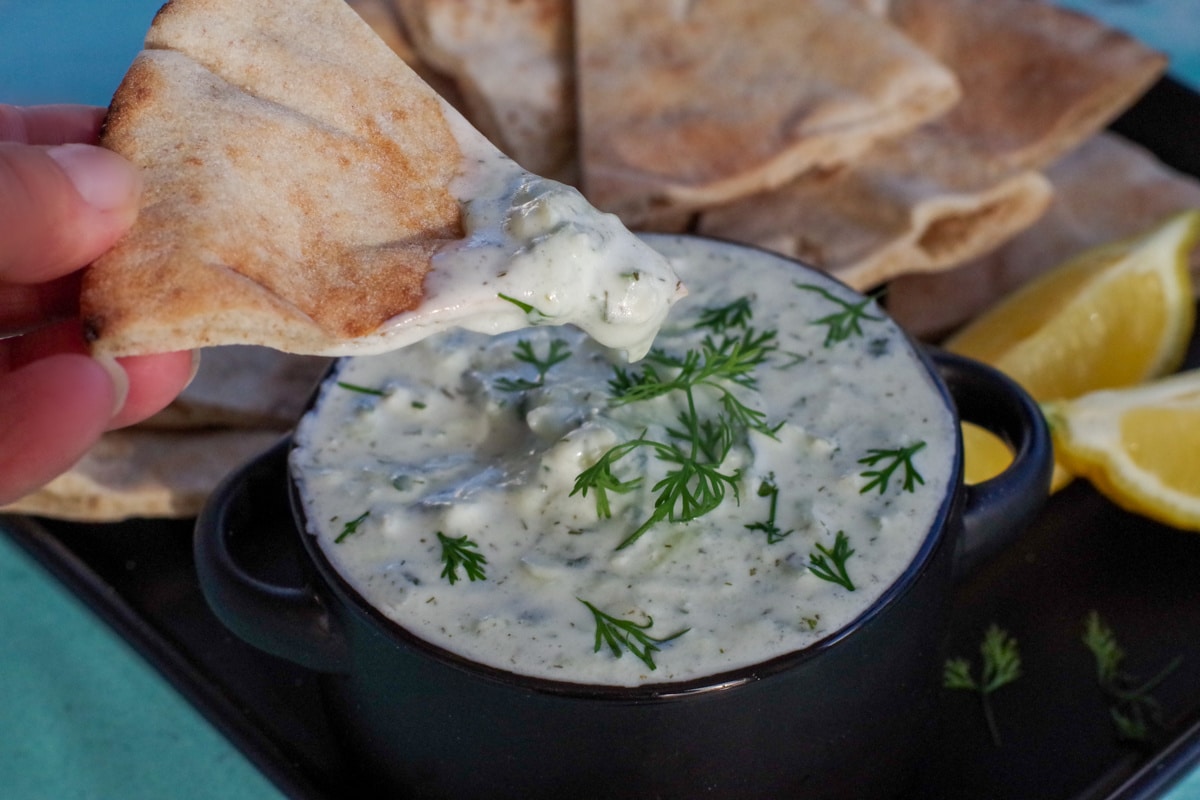 triangle of pita bread being dipped into tzatziki sauce in black bowl on black tray with pita bread in background and lemon wedges