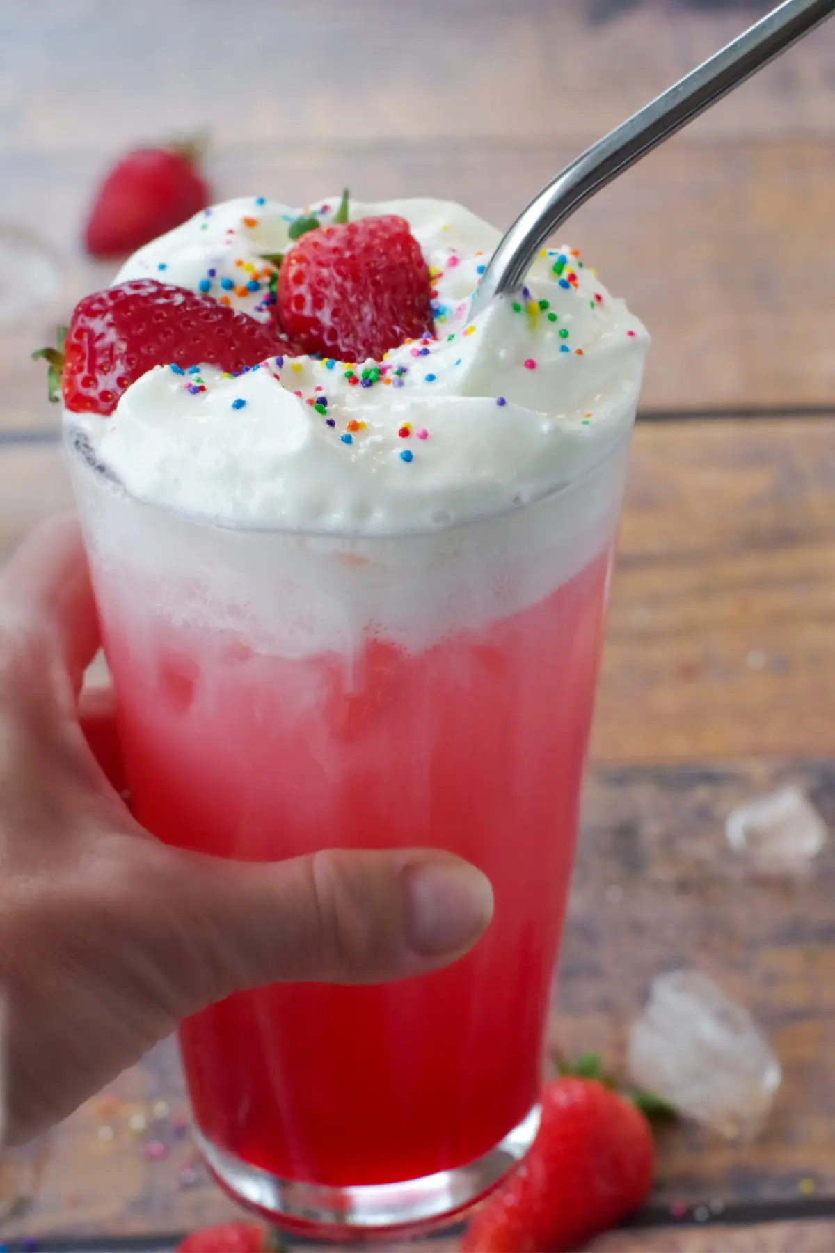 hand holding up Italian soda at an angle, with ice and strawberries in the background