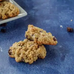 2 oatmeal raisin cookies on blue surface with plate of cookies, dish of raisins, jar of oats and scoop in the background