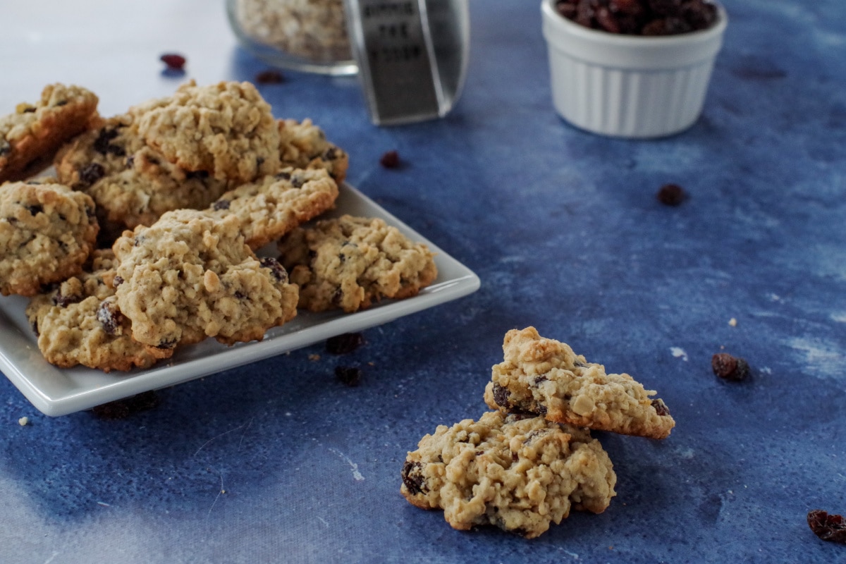 2 oatmeal raisin cookies on blue surface with plate of cookies, dish of raisins, jar of oats and scoop in the background