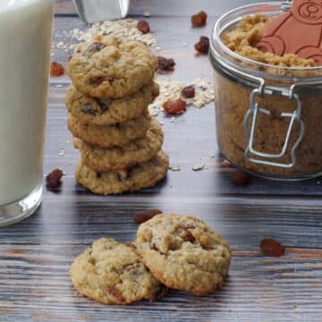 2 oatmeal raisin cookies (one resting partially on the other) in front of milk, a stack of more raisin cookies and jar of brown sugar