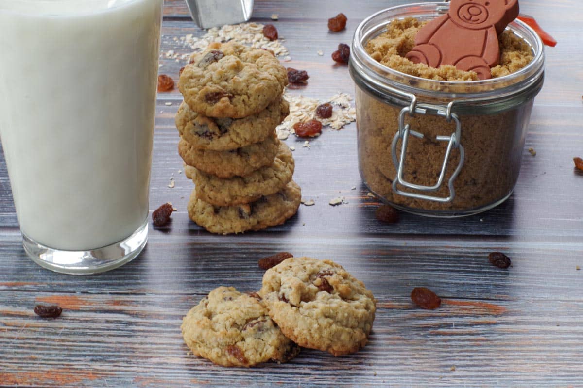 2 oatmeal raisin cookies (one resting partially on the other) in front of milk, a stack of more raisin cookies and jar of brown sugar