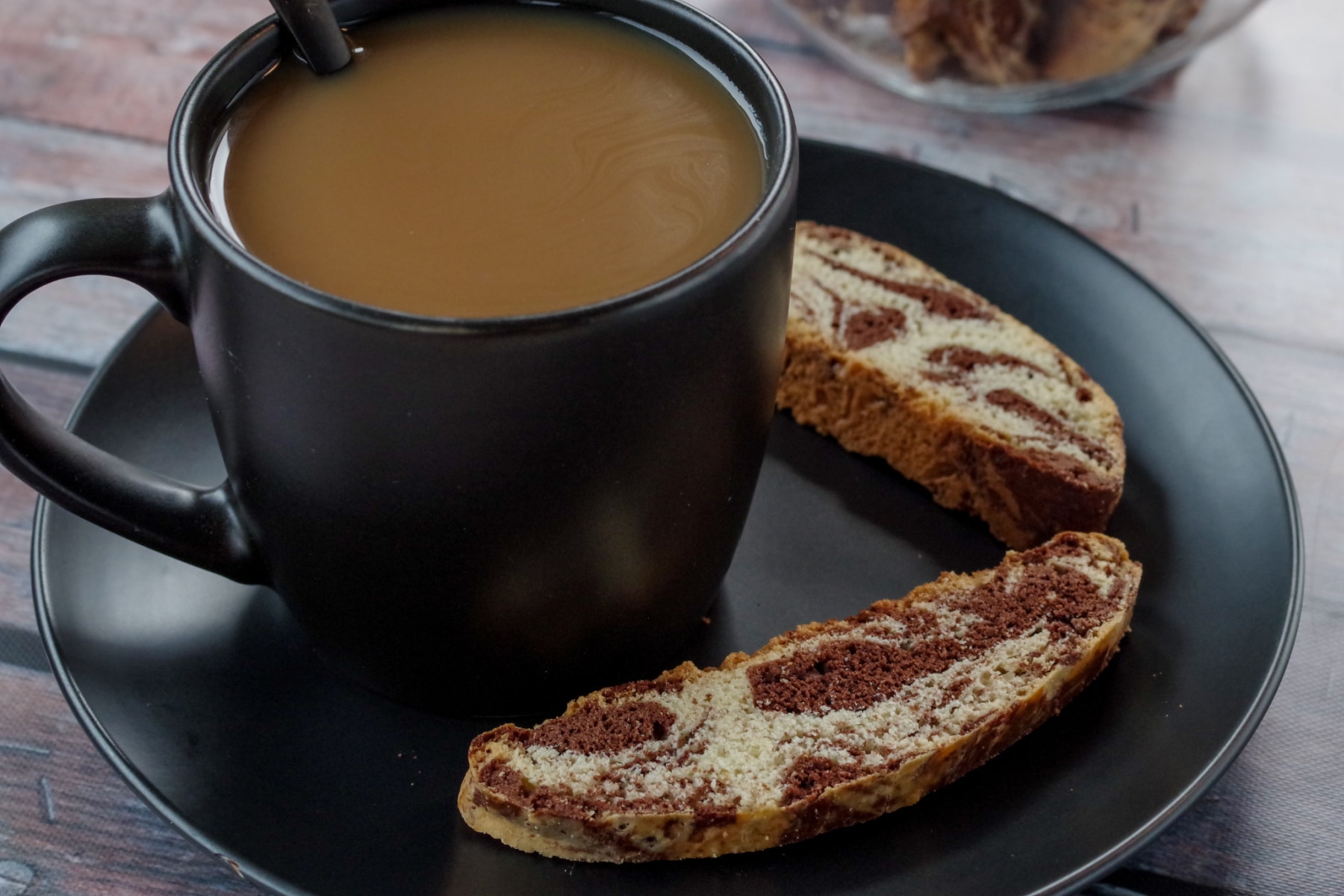 cup of coffee in a black mug on a black plate with 2 pieces of chocolate marble biscotti on the plate and a jar of biscotti in background