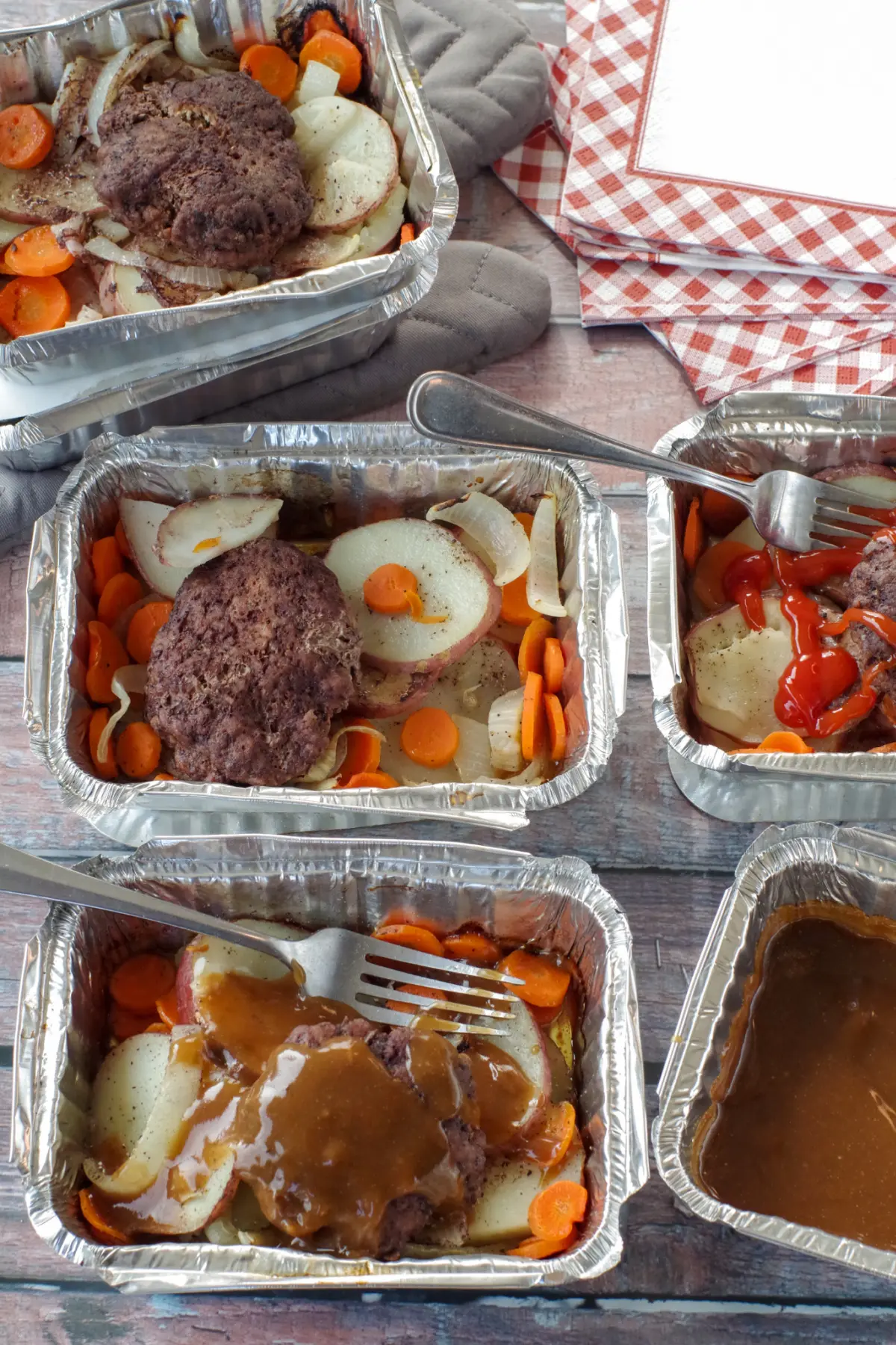 4 hamburger foil packets in aluminum foil containers with potatoes, carrots, onions and hamburgers, on wooden surface