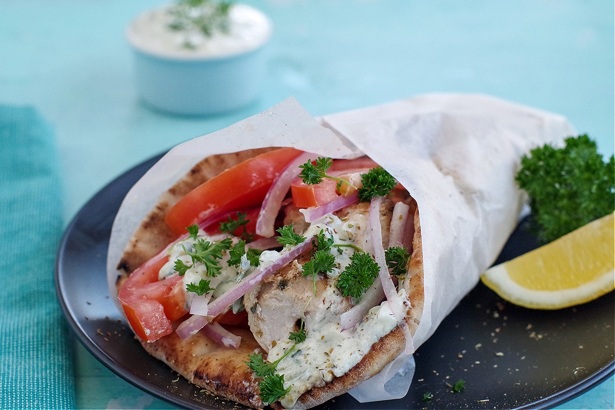 Greek chicken souvlaki wrapped in pita with tomatoes, red onion, tzatziki and parsley, wrapped in paper on black plate