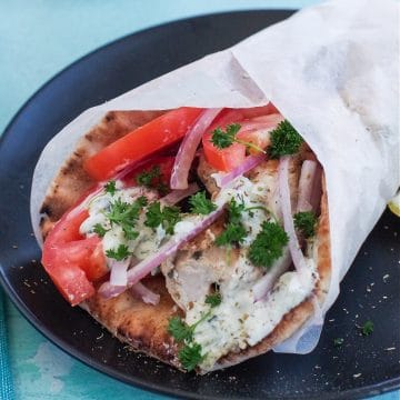 Greek chicken souvlaki wrapped in pita with tomatoes, red onion, tzatziki and parsley, wrapped in paper on black plate