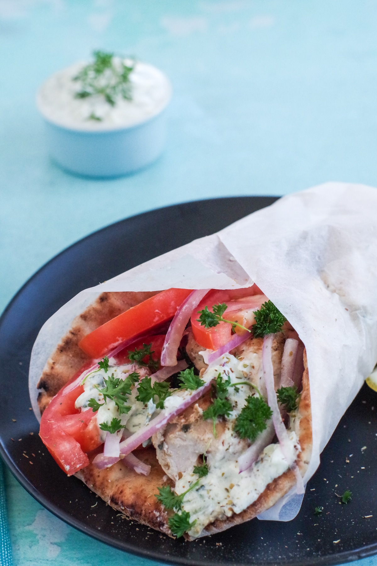 Greek chicken souvlaki wrapped in pita with tomatoes, red onion, tzatziki and parsley, wrapped in paper on black plate