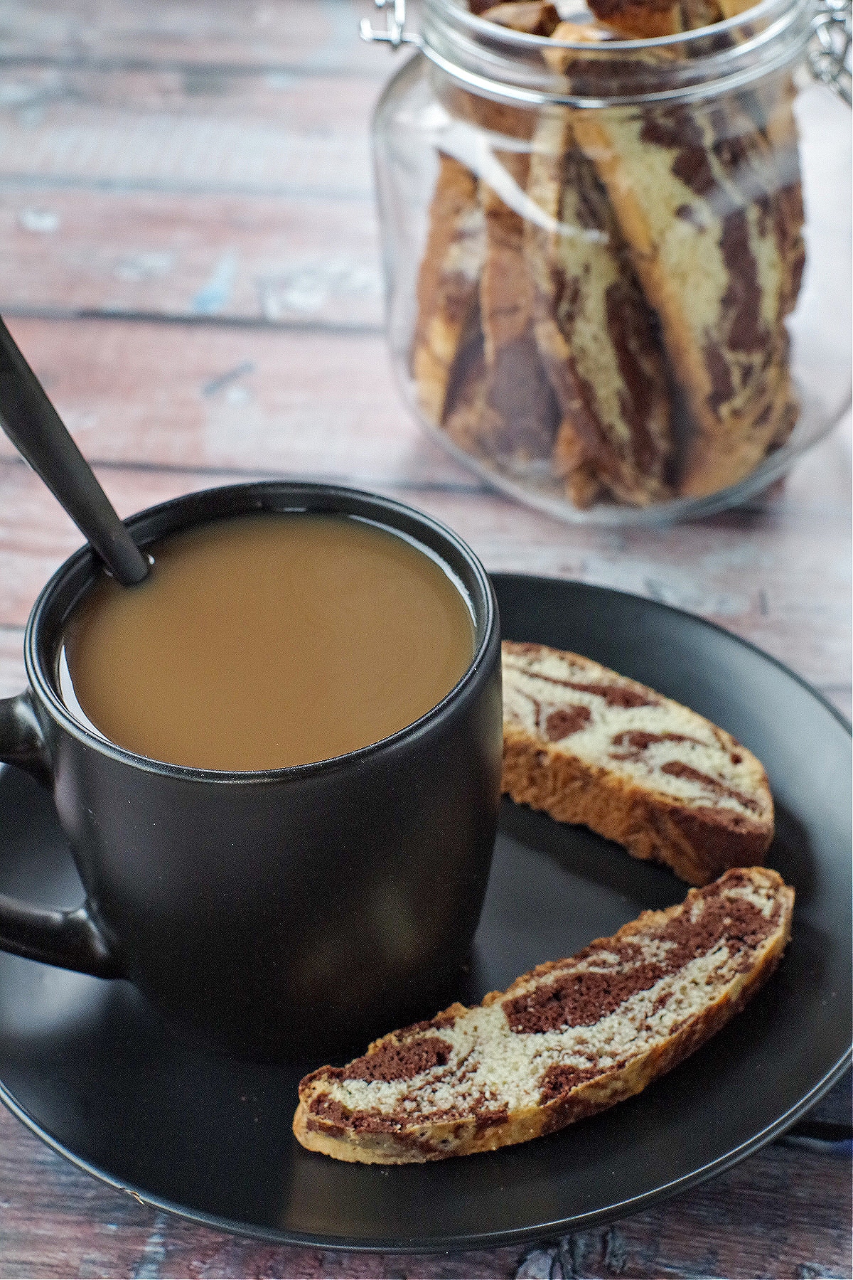 cup of coffee in a black mug on a black plate with 2 pieces of chocolate marble biscotti on the plate and a jar of biscotti in background