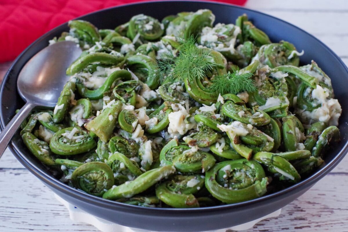 Cheesy Sauteed fiddleheads in a black bowl with a red oven mitt in the background