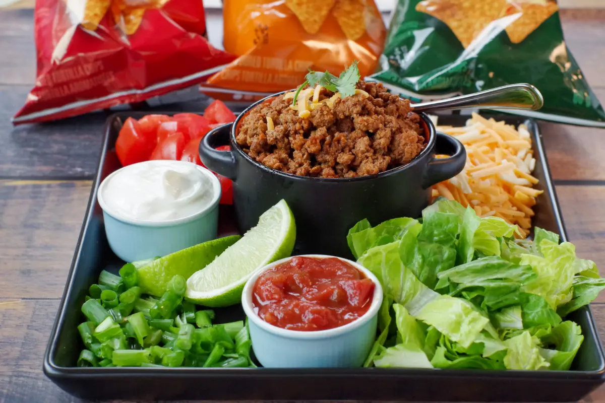 black tray with fixings (lettuce, salsa, sour cream, taco meat, lettuce, cheese, green onions and tomatoes, with bags of tortilla chips in the backgroun