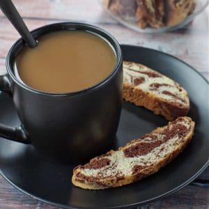 cup of coffee in a black mug on a black plate with 2 pieces of chocolate marble biscotti on the plate and a jar of biscotti in background