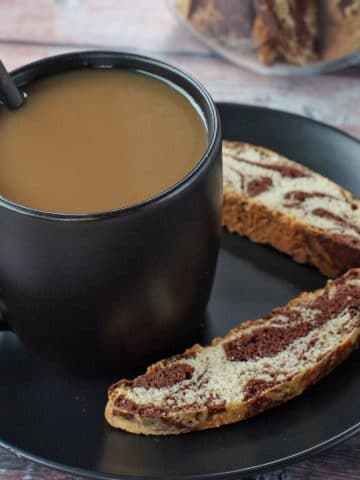 cup of coffee in a black mug on a black plate with 2 pieces of chocolate marble biscotti on the plate and a jar of biscotti in background