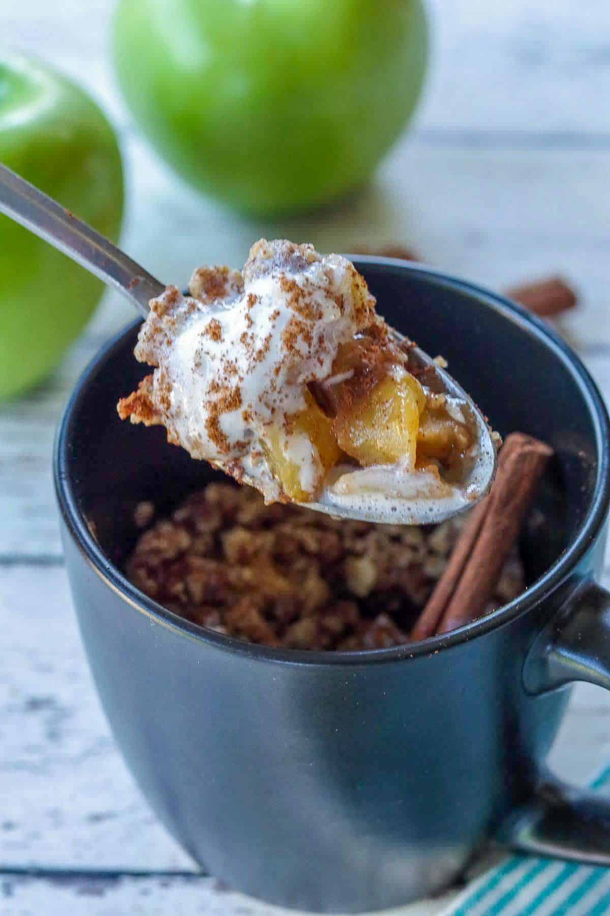 spoon with apple crisp being held up above a black mug apple crisp with 2 green apples and cinnamon sticks in the background