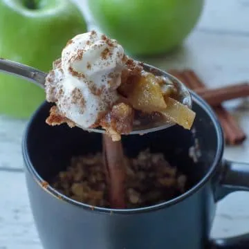 spoon with apple crisp being held up above a black mug apple crisp with 2 green apples and cinnamon sticks in the background