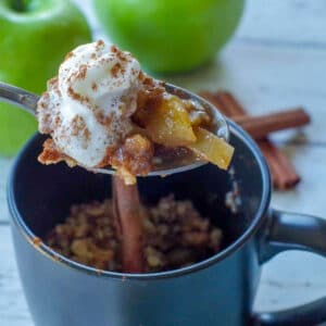 spoon with apple crisp being held up above a black mug apple crisp with 2 green apples and cinnamon sticks in the background