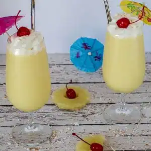 2 health pina colada drinks on a white faux wood surface, with pineapple, maraschino cherries and umbrellas
