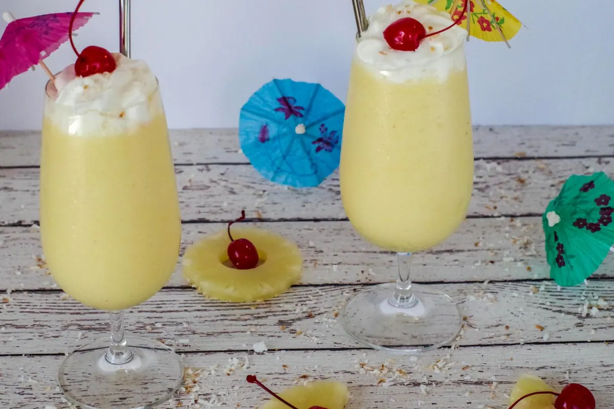 2 health pina colada drinks on a white faux wood surface, with pineapple, maraschino cherries and umbrellas