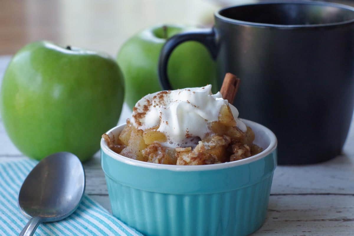 mug apple crisp in a blue dish with black mug and 2 green apples in the background