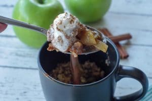 spoon with apple crisp being held up above a black mug apple crisp with 2 green apples and cinnamon sticks in the background