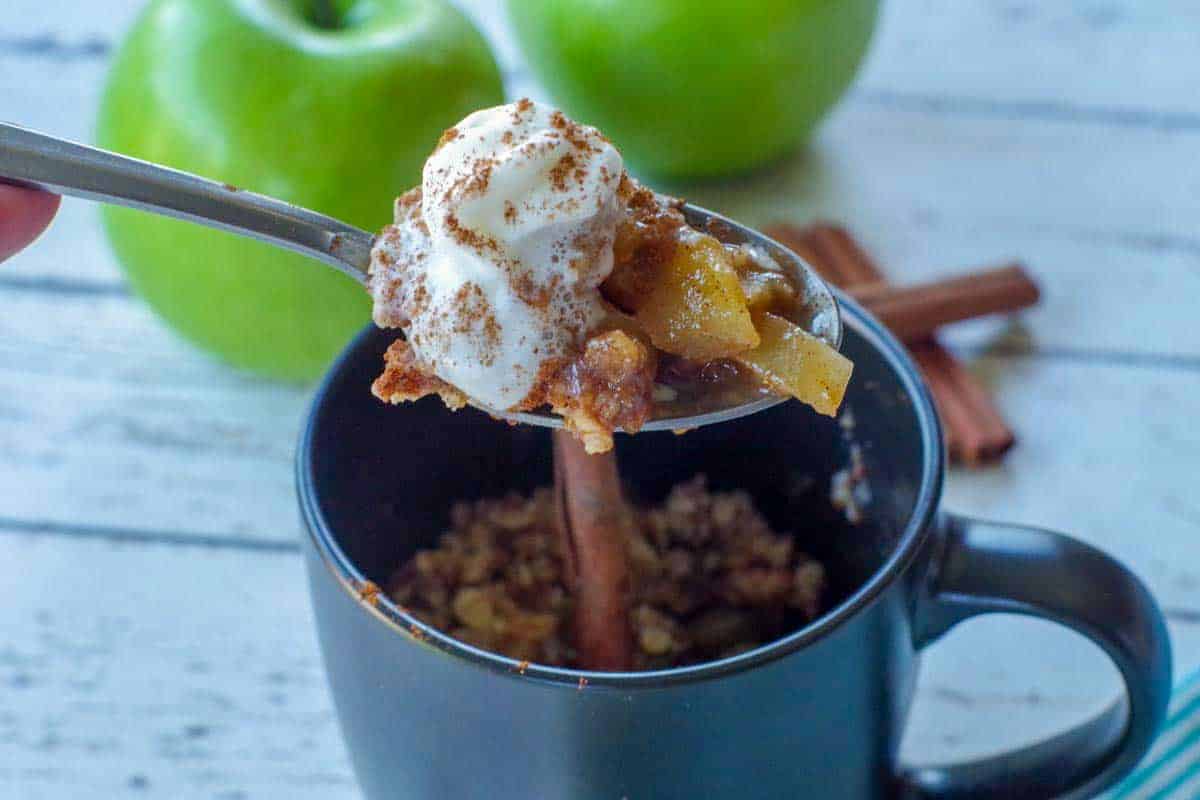 spoon with apple crisp being held up above a black mug apple crisp with 2 green apples and cinnamon sticks in the background