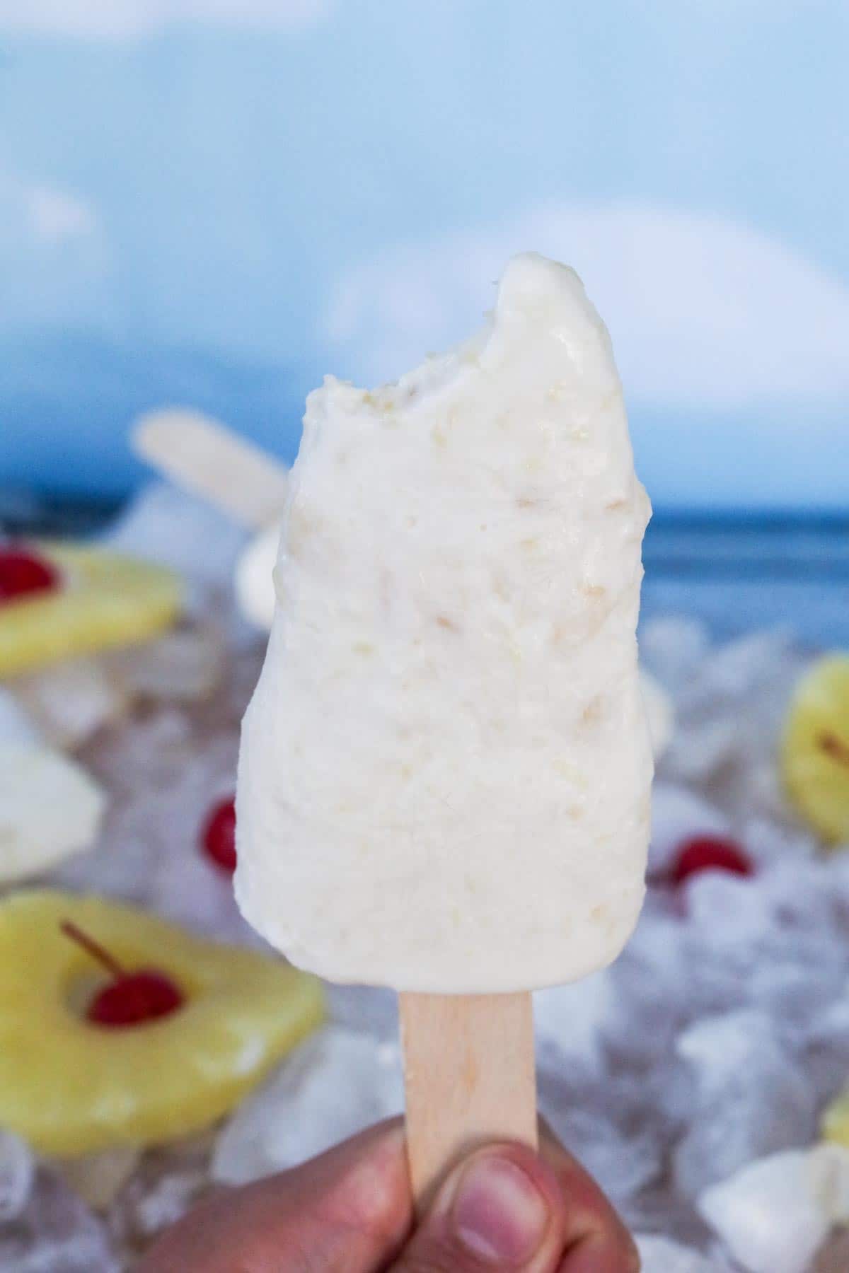 pina colada popsicle, with a bite taken out of it, being held up by hand and ice, pineapple rings, cherries and more popsicles in the background