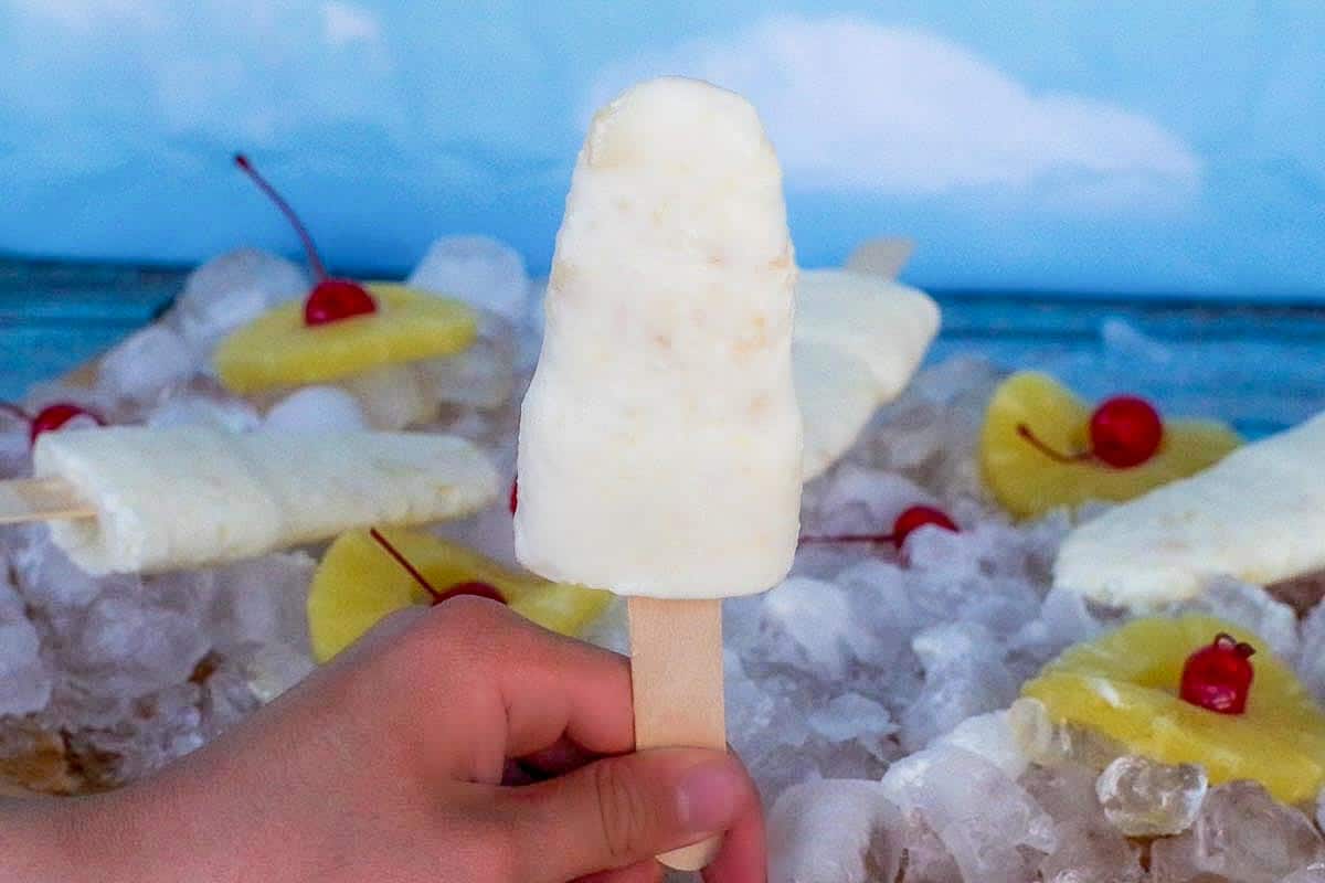 pina colada popsicle being held up by hand and ice, pineapple rings, cherries and more popsicles in the background