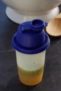 salad dressing in a Tupperware salad shaker with wooden spoon and white bowl in the background