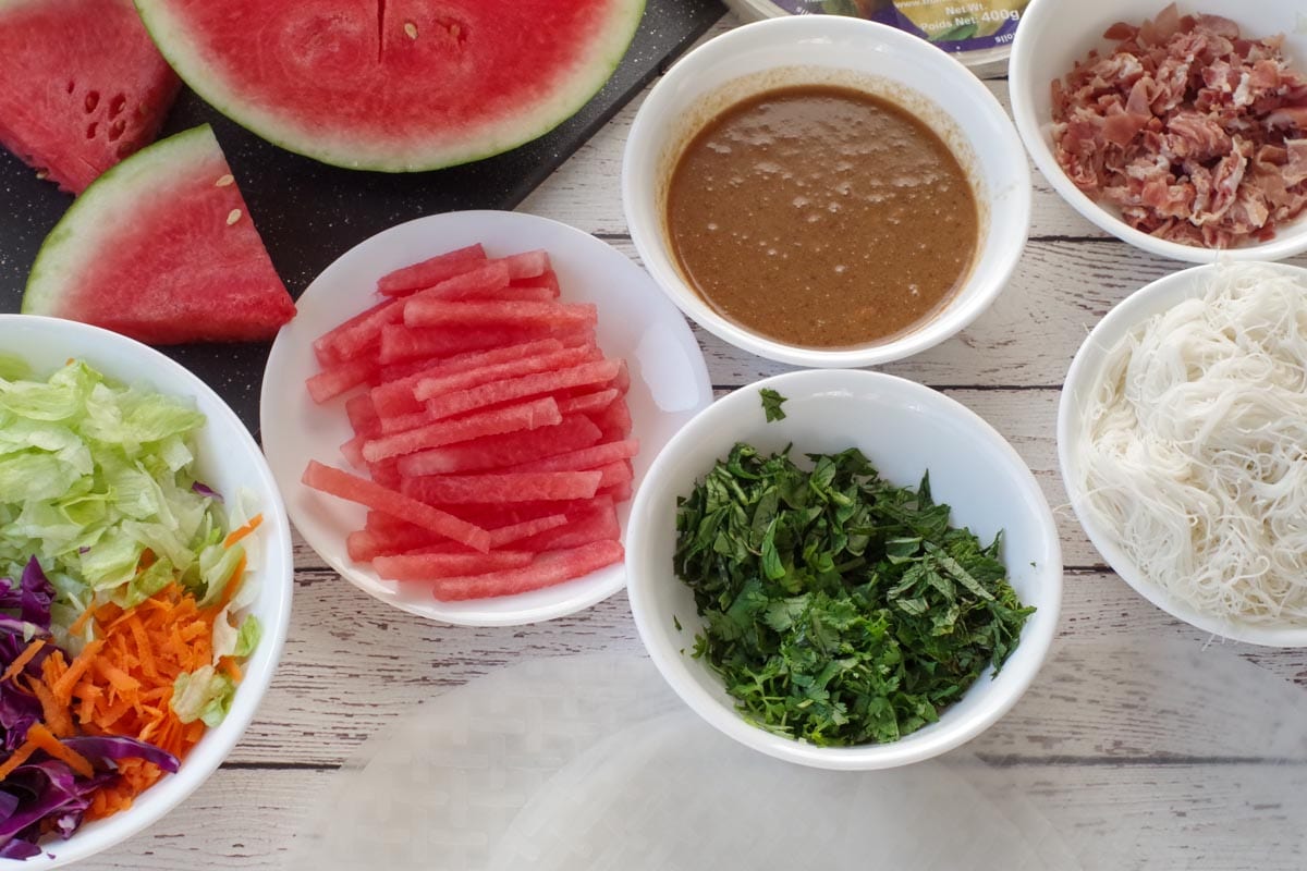 ingredients for watermelon prosciutto salad rolls in bowls and plates, with watermelon in background and rice paper in foreground