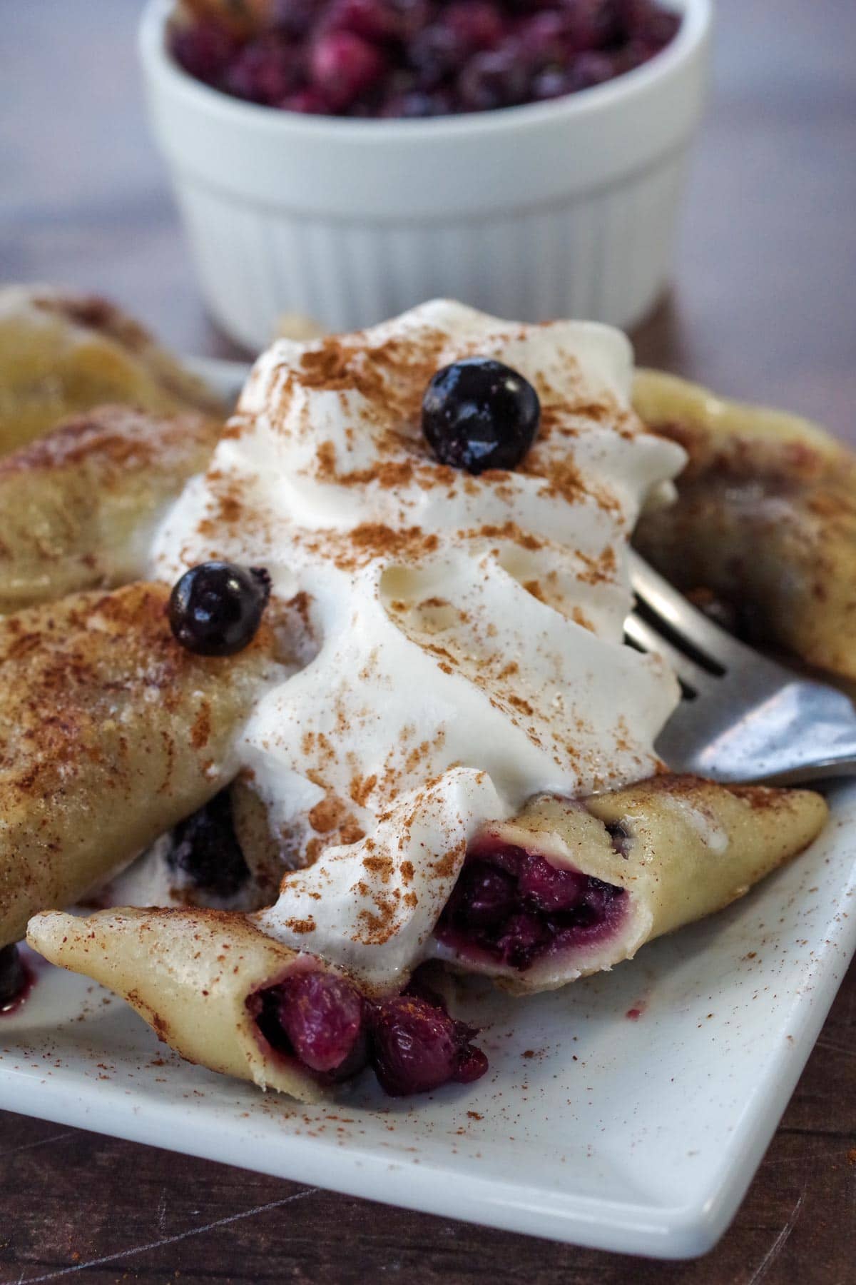 Sasktoon berry perogies with cinnamon and whipped cream, on a white plate with a dish of Saskatoon berry filling in the background