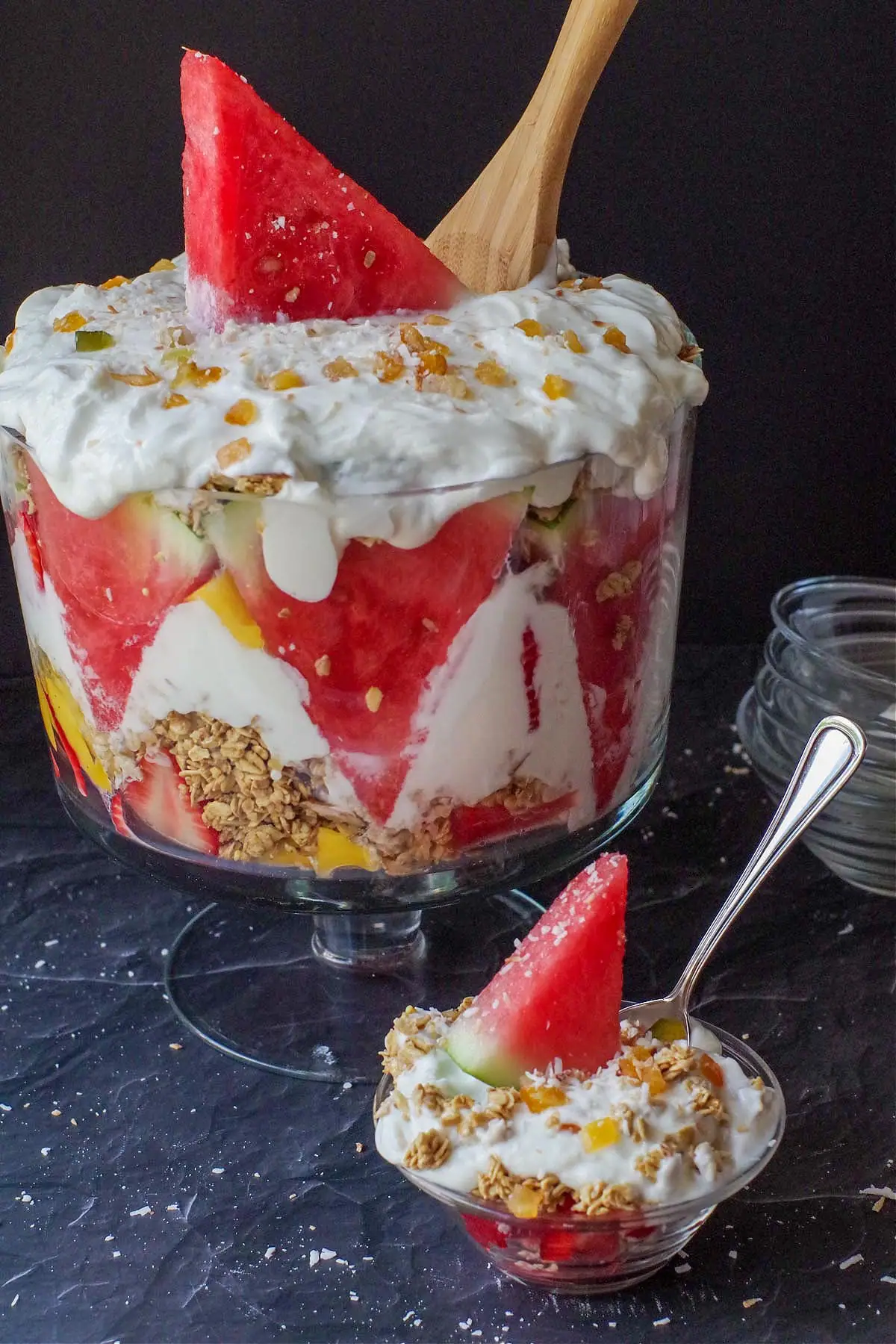 watermelon breakfast parfait in a glass bowl with spoon sticking out and large trifle bowl full of watermelon granola trifle in background