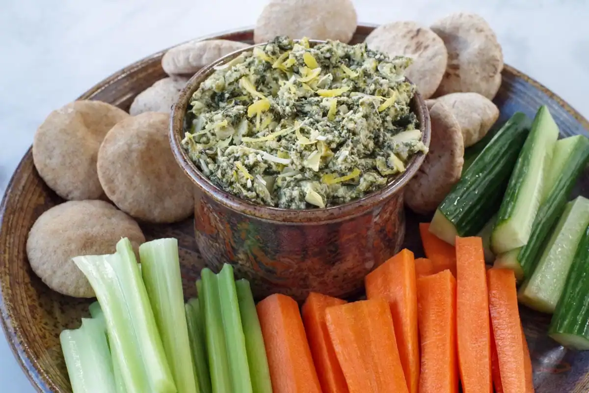 healthy spinach artichoke dip in a ceramic bowl on a platter with vegetables and mini pita breads
