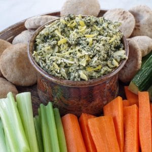 healthy spinach artichoke dip in a ceramic bowl on a platter with vegetables and mini pita breads