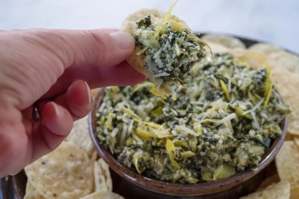spinach artichoke dip being held up on a chip over a platter of more dip and tortilla chips