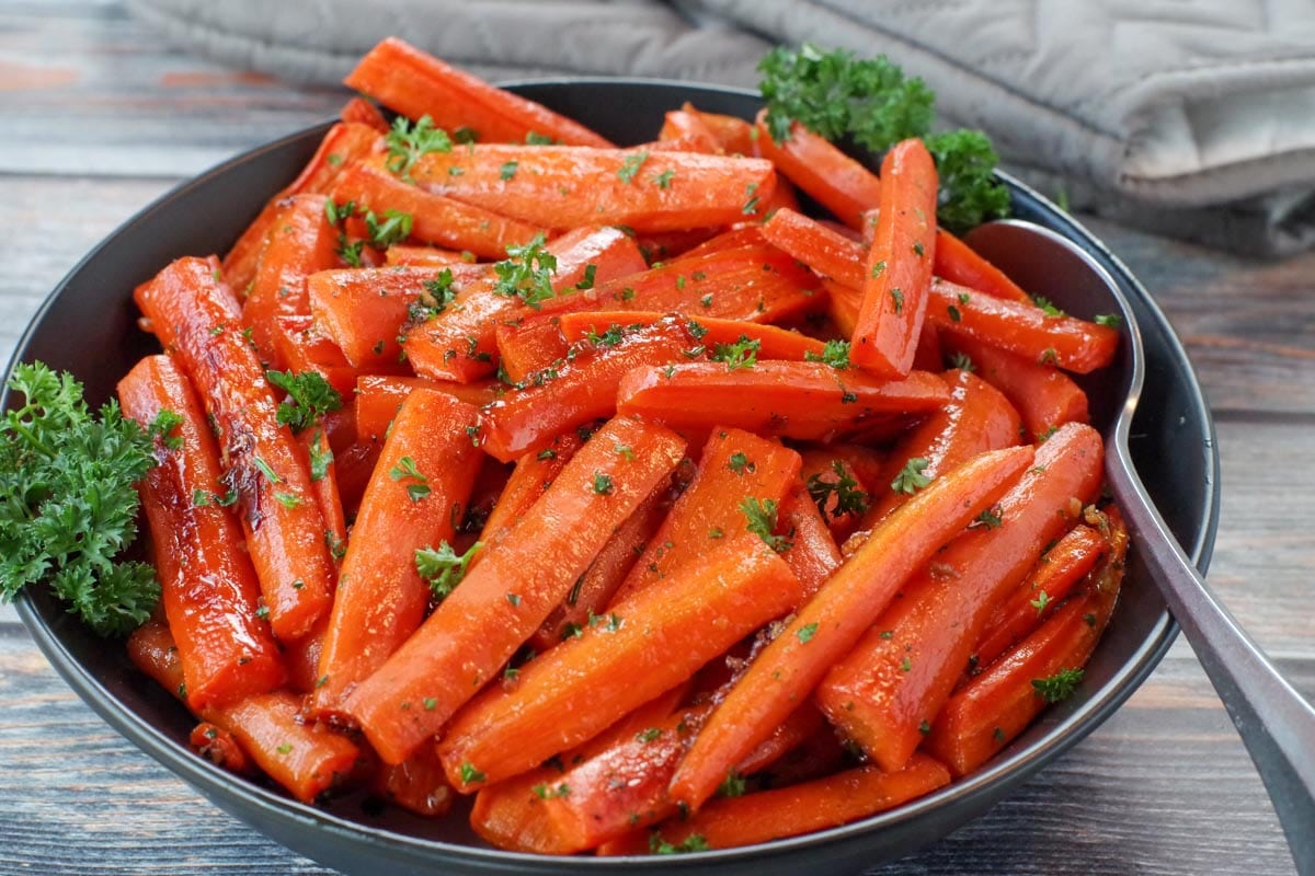 healthy roasted carrots with brown sugar in a black bowl, with a spoon and grey oven mitts in the background